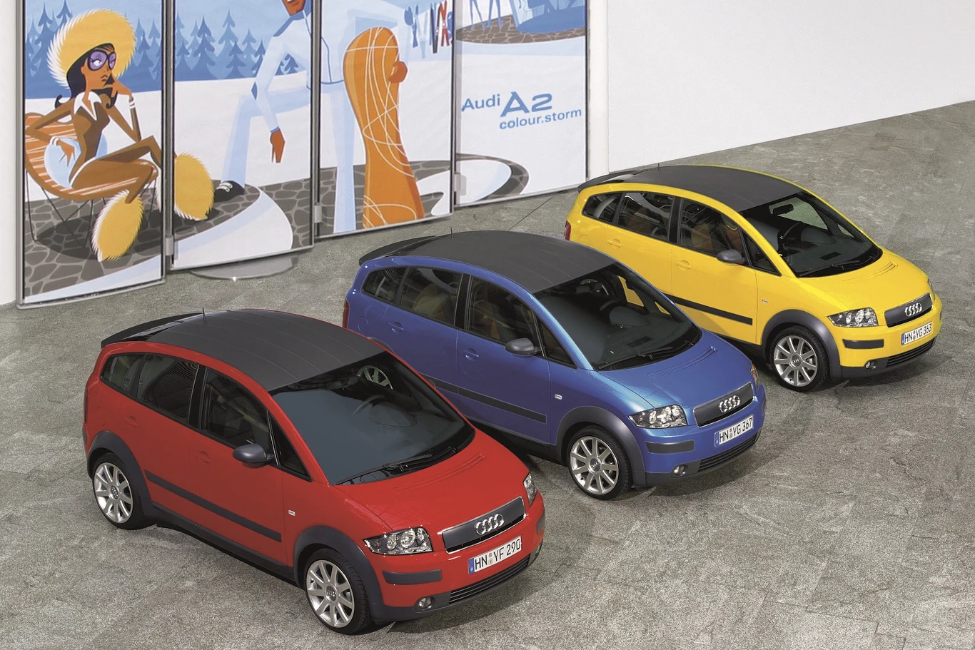 Trzy samochody Audi A2 w różnych kolorach: czerwonym, niebieskim i żółtym, ustawione obok siebie na placu; każdy z nich ma czarny dach, a w tle widoczny jest kolorowy plakat z zimowym motywem i napisem 'Audi A2 colour storm'.