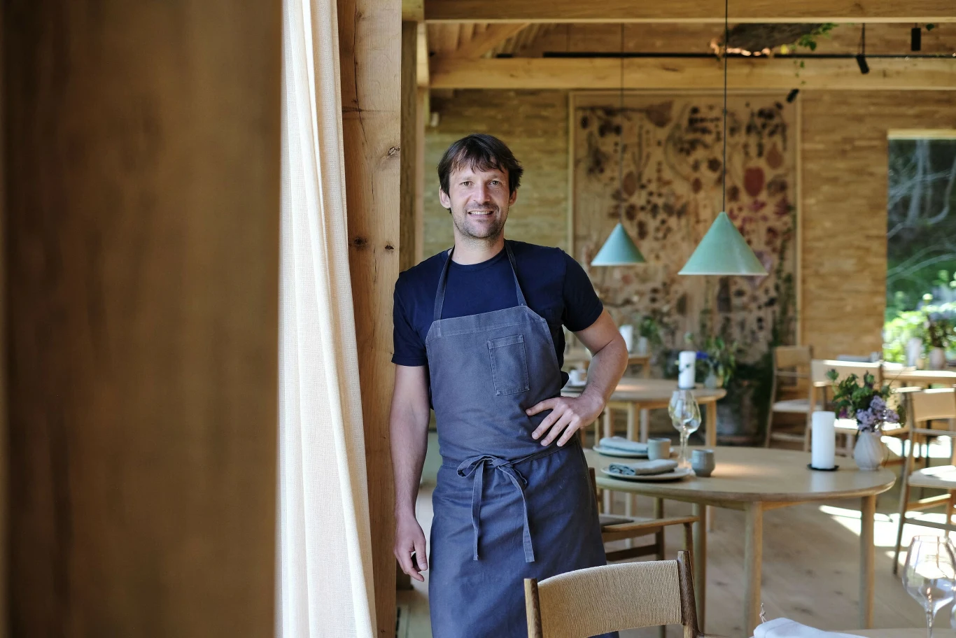 uśmiechnięty mężczyzna w szarym fartuchu stoi w przytulnie urządzonej restauracji z drewnianymi meblami, dekoracyjnymi lampami i kwiatami na stołach