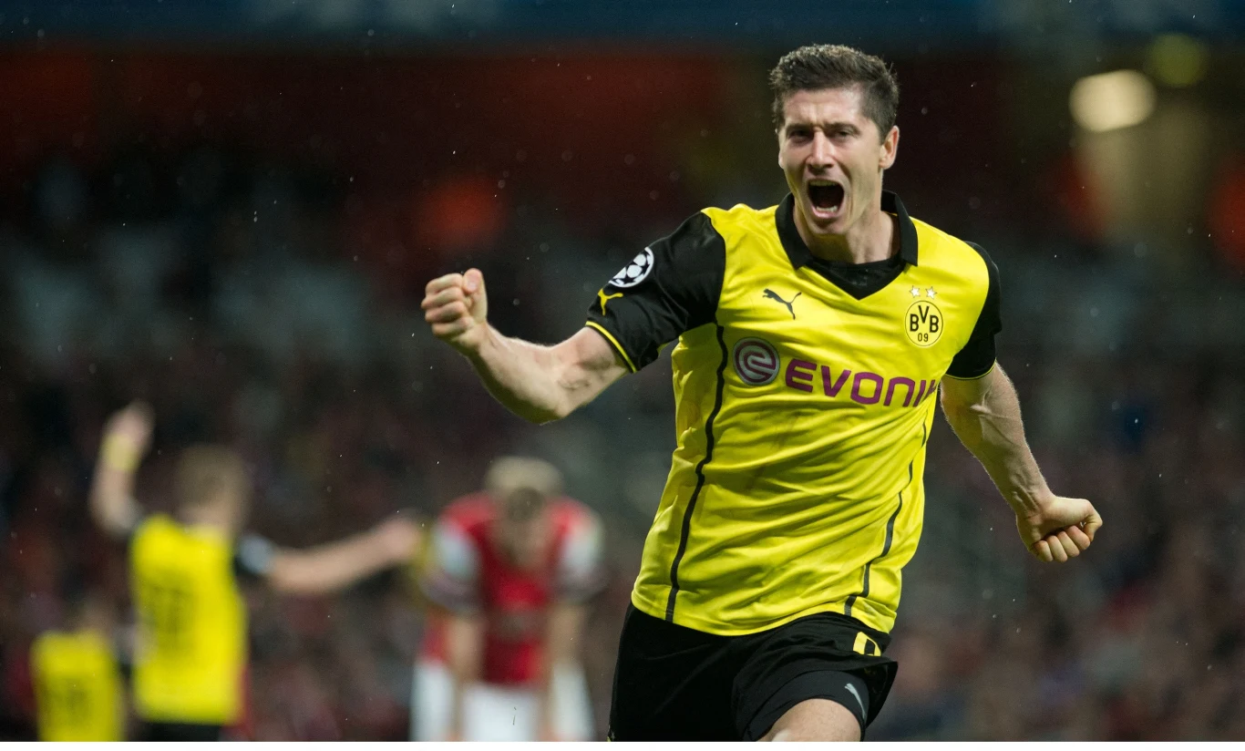 Piłkarz Borussii Dortmund w żółto-czarnym stroju biegnie z uniesioną pięścią, wyrażając silne emocje po zdobyciu gola podczas meczu, na tle rozmazanych zawodników drużyny przeciwnej.