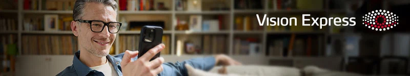 Mężczyzna w okularach trzymający smartfon i uśmiechający się, siedzący na kanapie w jasnym, nowoczesnym wnętrzu z półkami na książki w tle, obok logo Vision Express.