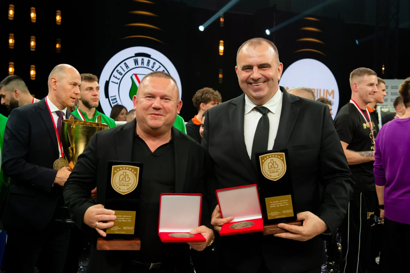 Dwóch uśmiechniętych mężczyzn w garniturach trzyma w dłoniach statuetki i otwarte pudełka z medalami, w tle grupa ludzi w sportowych strojach oraz duże logo klubu sportowego.