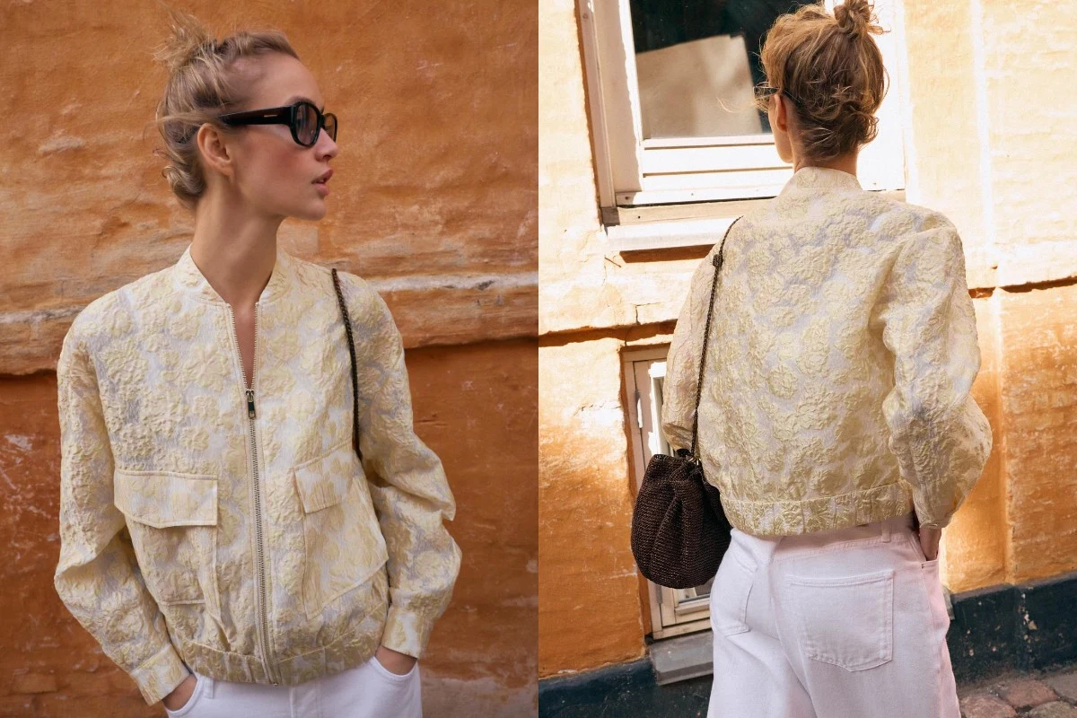 Kobieta w jasnej, wzorzystej bomberce i białych spodniach stoi na tle pomarańczowej ściany, ma spięte włosy, nosi duże, ciemne okulary przeciwsłoneczne i brązową torebkę na ramieniu.