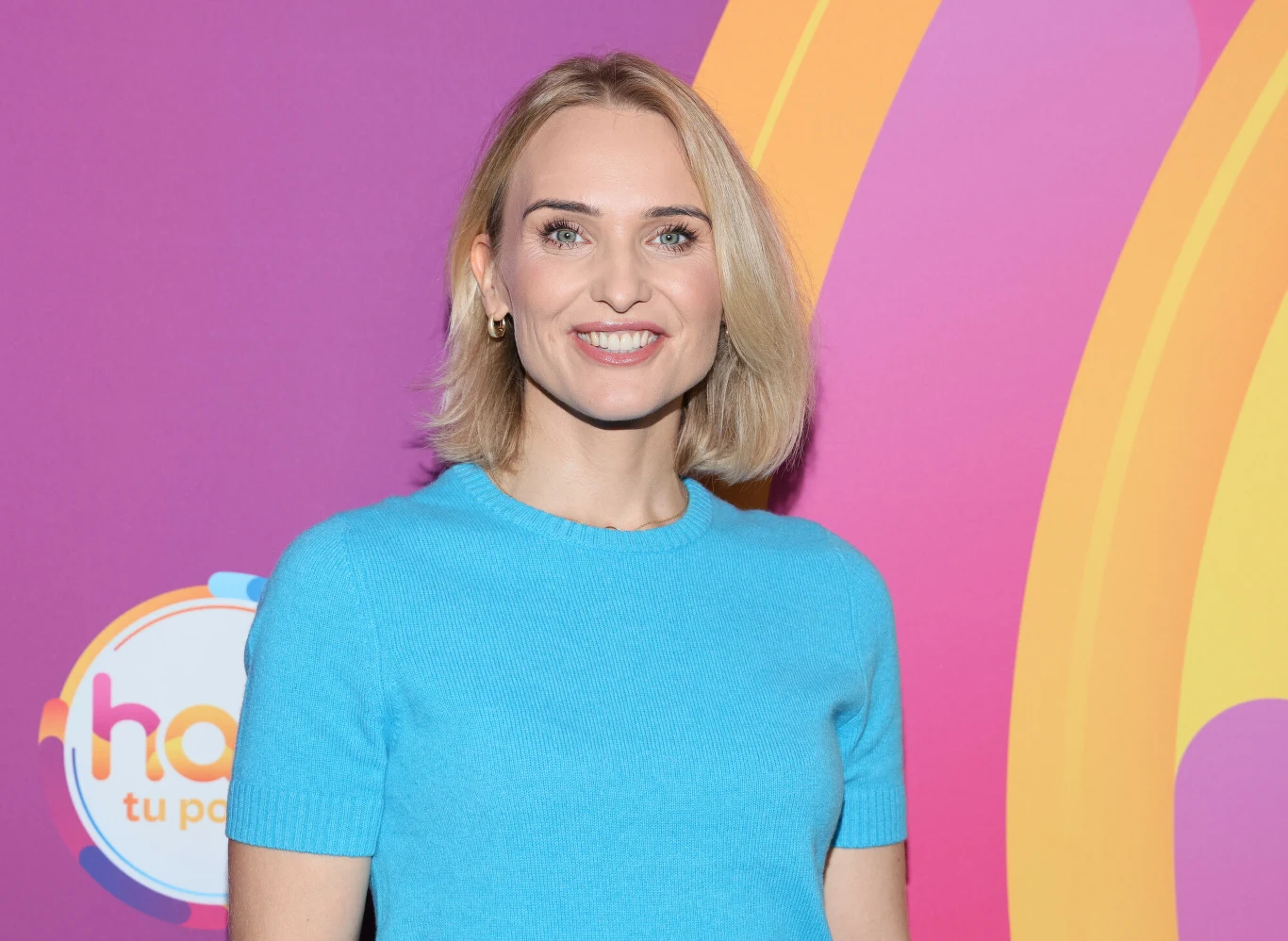 Kobieta z blond włosami, ubrana w błękitny sweter, stoi na tle fioletowo-żółtej grafiki z kolorowymi napisami.