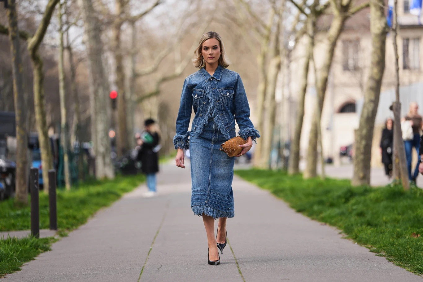 kobieta idąca środkiem miejskiego chodnika, ubrana w dżinsową kurtkę i spódnicę midi z frędzlami, trzymająca brązową torebkę i nosząca czarne szpilki; w tle wiosenne drzewa i niewyraźne sylwetki przechodniów