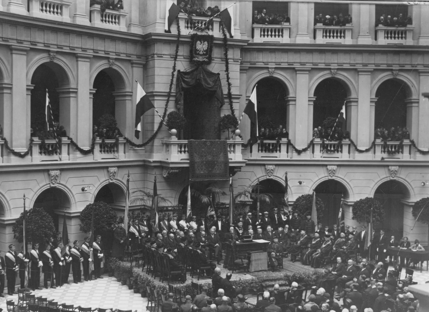 Uroczysta ceremonia w okazałym, zabytkowym dziedzińcu otoczonym arkadami, z licznymi uczestnikami w oficjalnych strojach zgromadzonymi wokół podwyższenia, na którym znajdują się najważniejsze postacie, a na balkonach i przy ścianach obserwuje scena wie...