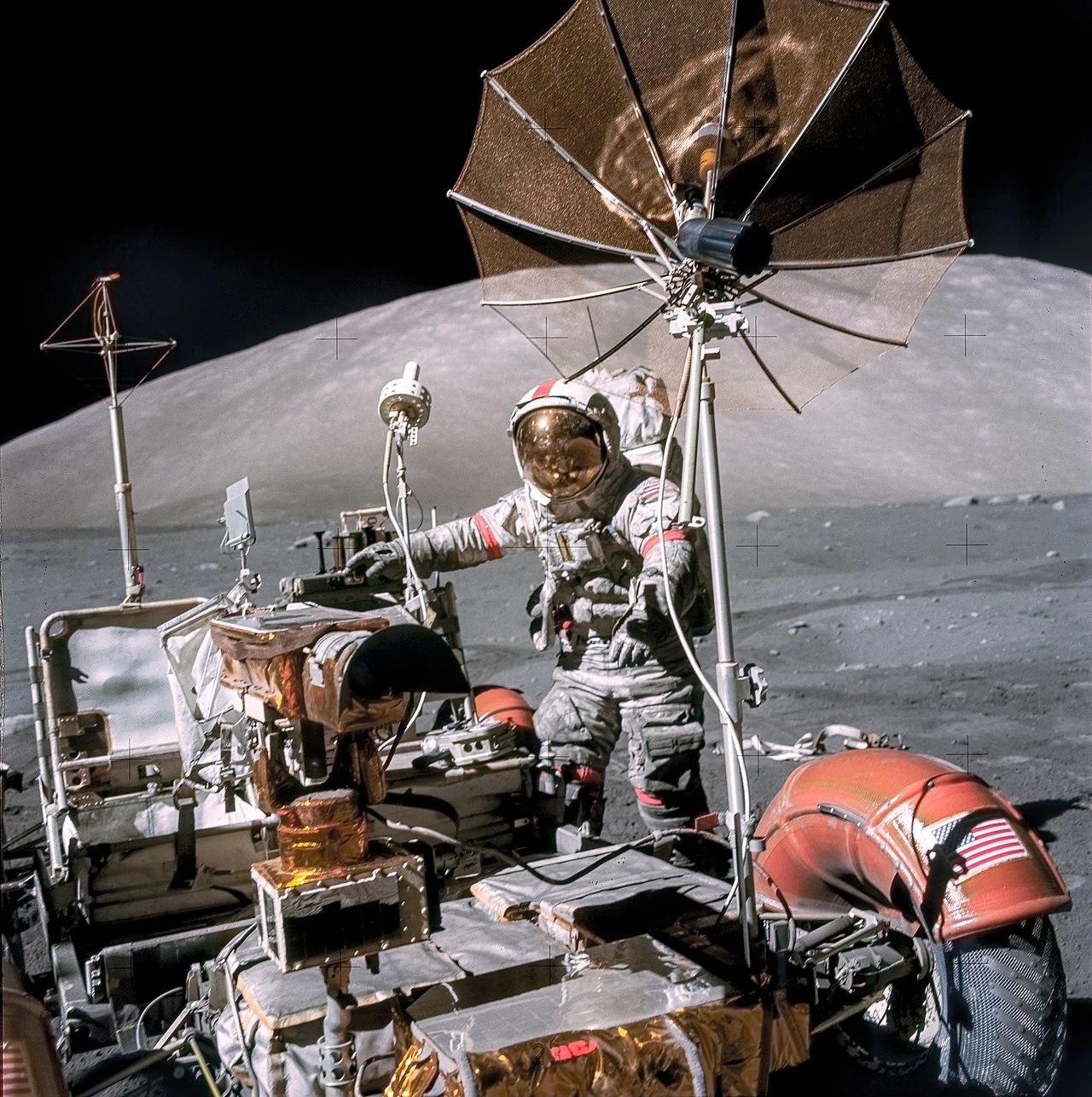 astronauta w skafandrze kosmicznym operujący łazikiem księżycowym wyposażonym w antenę paraboliczną na powierzchni Księżyca, w tle widoczny szary, pagórkowaty krajobraz
