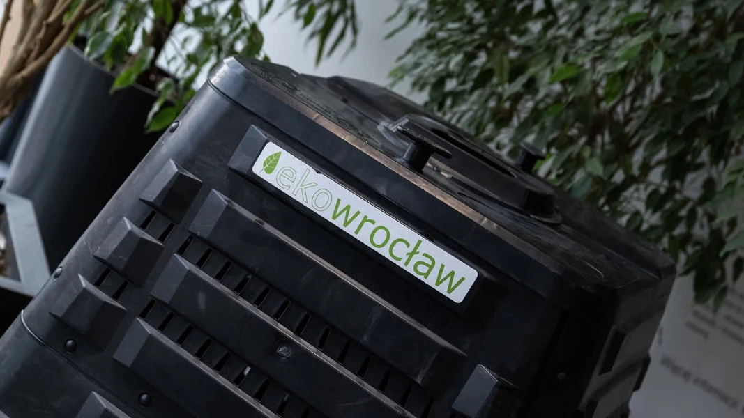 czarny pojemnik do kompostowania z naklejką „ekoWrocław”, otoczony przez doniczkowe rośliny