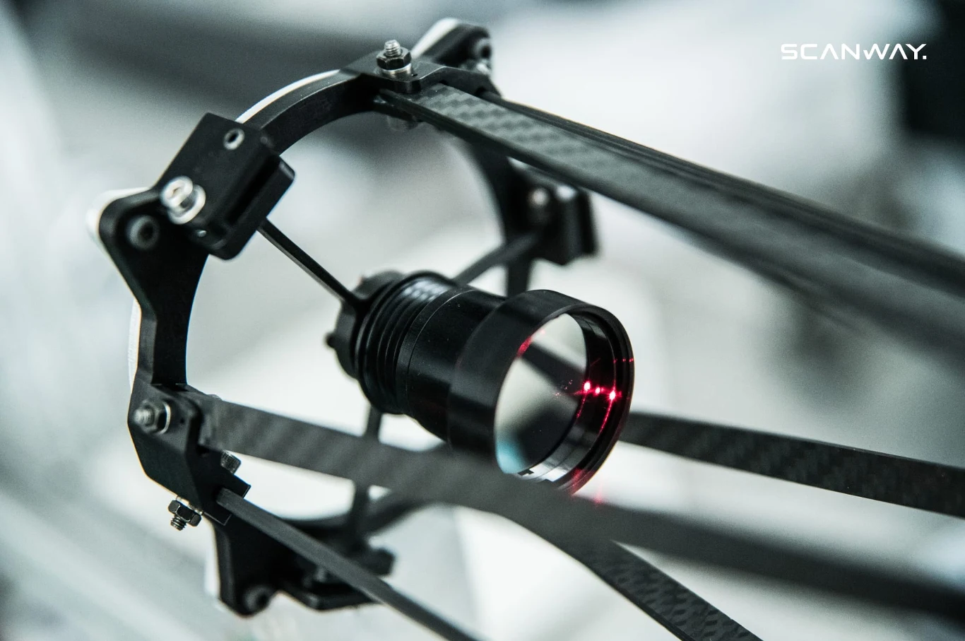 Obiektyw optyczny zamocowany w metalowej ramie z elementami z włókna węglowego, przez który przechodzi wiązka czerwonego światła.