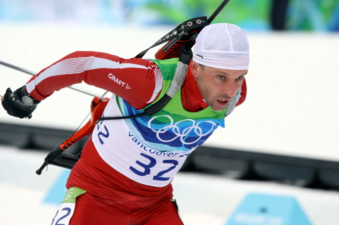 biathlonista w czerwonym stroju sportowym, jadący na nartach biegowych z karabinem na plecach, z numerem startowym 32, w trakcie zawodów olimpijskich