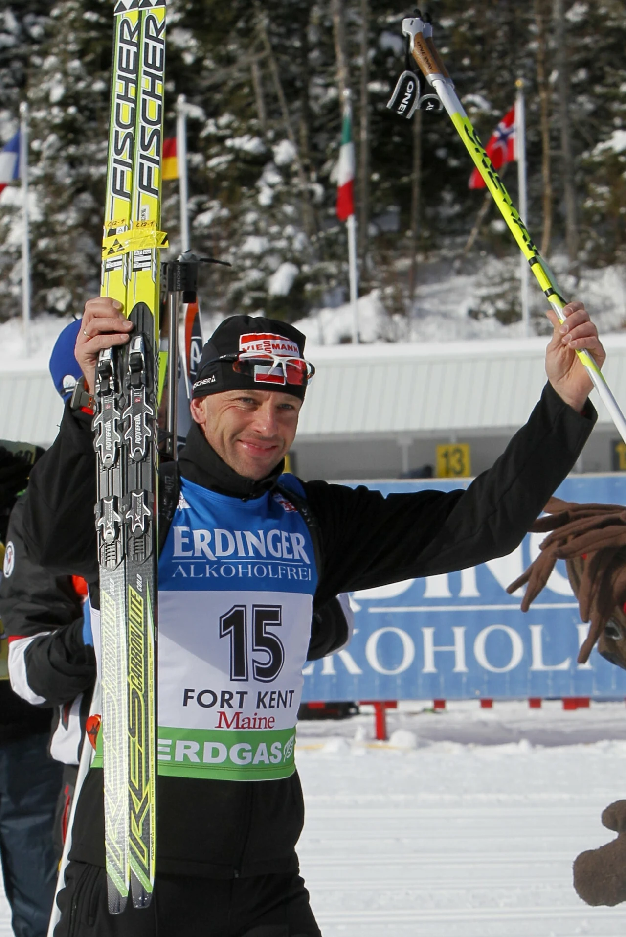 biatlonista w sportowym stroju z nartami w jednej ręce i kijem narciarskim uniesionym w drugiej, z uśmiechem na twarzy po zakończonym wyścigu, w tle zimowy krajobraz oraz powiewające flagi państw