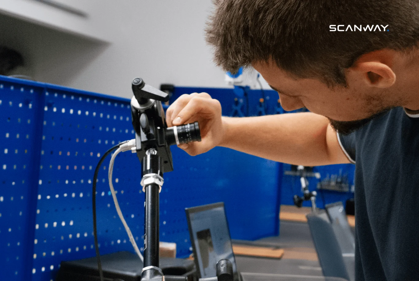 mężczyzna reguluje urządzenie optyczne zamocowane na statywie, w tle niebieski panel warsztatowy oraz stanowisko robocze z komputerem