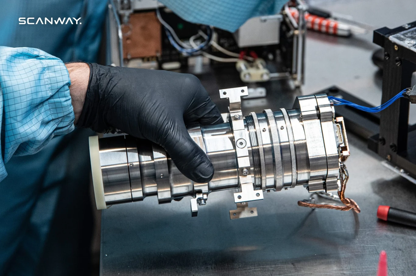 osoba w niebieskim fartuchu ochronnym i czarnych rękawiczkach trzyma precyzyjne urządzenie optyczne wykonane z metalu, prawdopodobnie część systemu optycznego lub kamery przemysłowej, na stole widać narzędzia oraz elementy elektroniczne