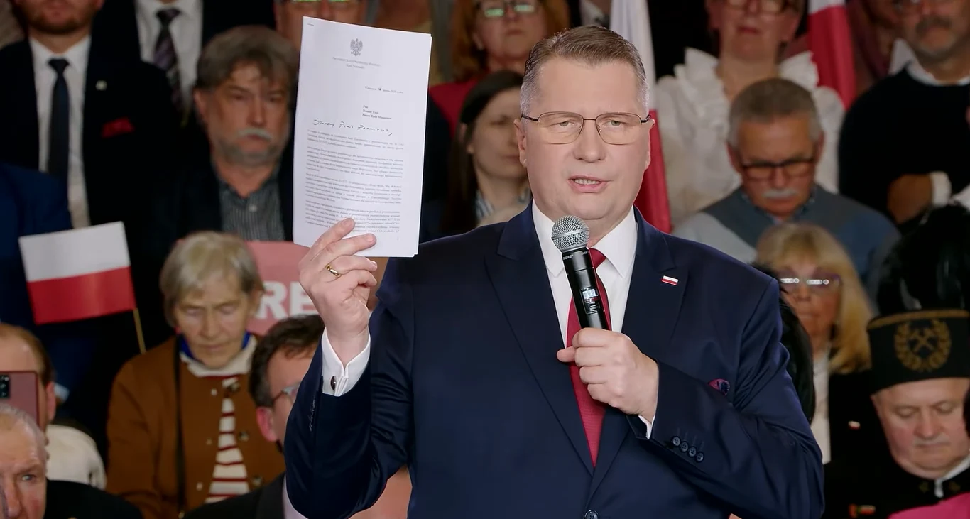 Mężczyzna w garniturze i okularach trzyma w jednej ręce mikrofon, a w drugiej oficjalny dokument, stojąc przed grupą osób z polskimi flagami w tle.