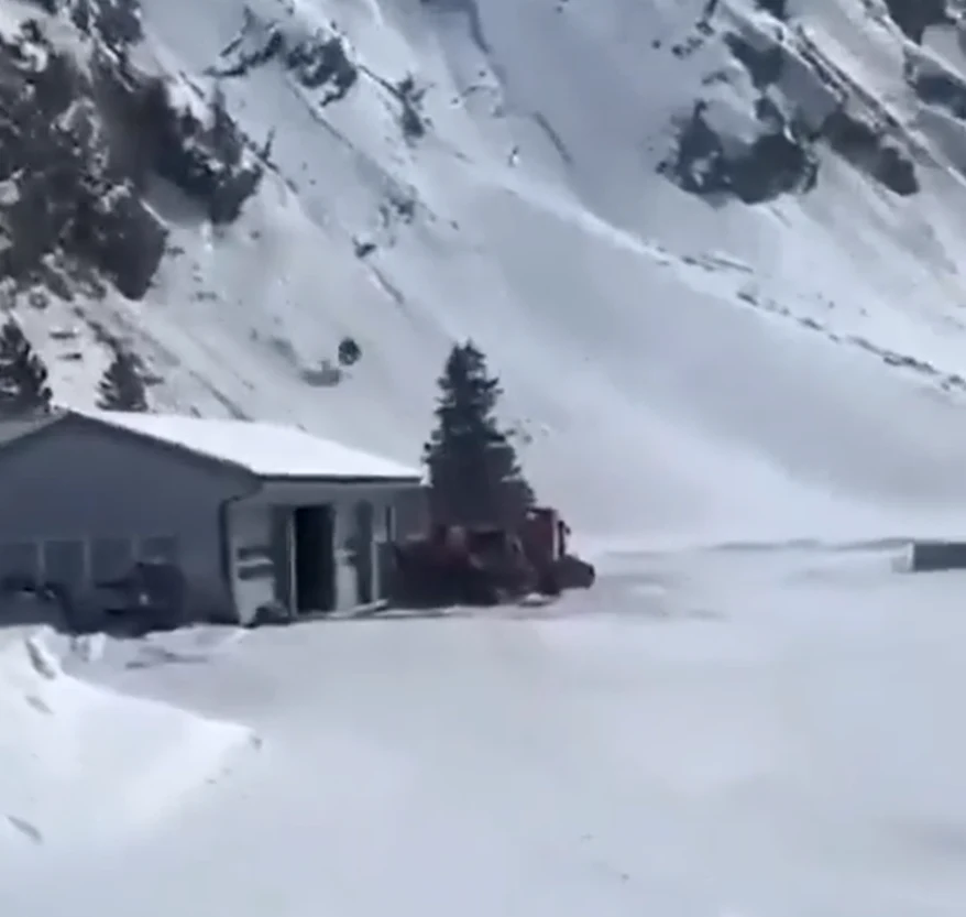 Samotny budynek w otoczeniu wysokich, ośnieżonych gór, obok którego stoi pojazd ciężarowy oraz pojedyncze drzewo iglaste; wszystko pokryte grubą warstwą śniegu.