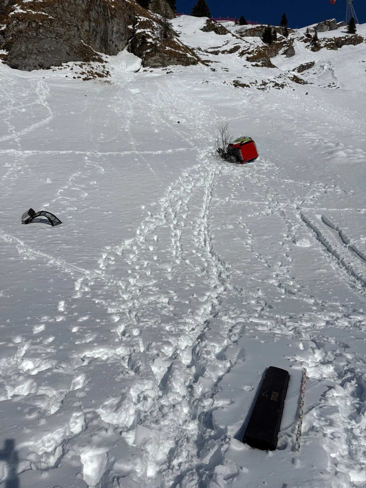 przewrócony ratrak na stromym, ośnieżonym zboczu, rozrzucone fragmenty maszyny oraz ślady pozostawione przez ludzi na śniegu