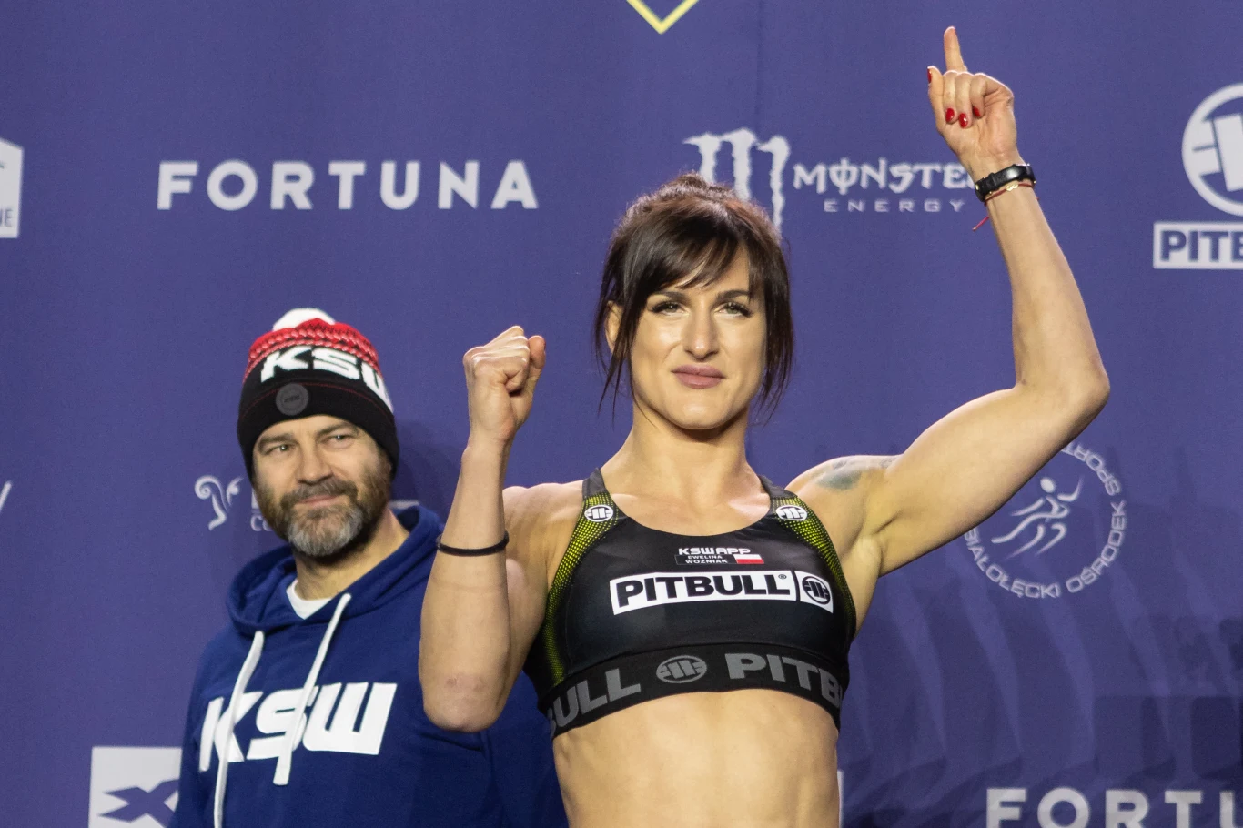 zawodniczka w sportowym stroju z uniesioną ręką, prezentująca umięśnioną sylwetkę, na tle ściany z logo sponsorów oraz mężczyzna w czapce i bluzie z logo KSW stojący obok