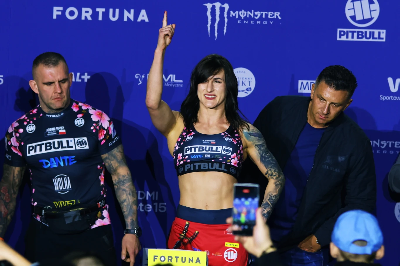 Kobieta w sportowym stroju unosi rękę z uniesionym palcem do góry, stojąc pomiędzy dwoma mężczyznami podczas wydarzenia sportowego na tle niebieskiej ściany z logotypami sponsorów.