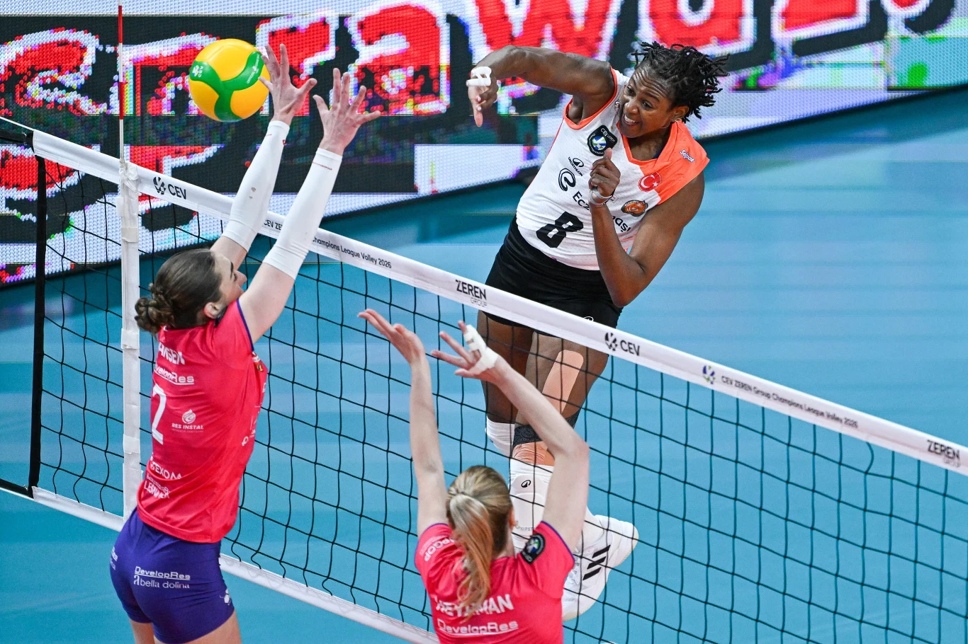 siatkarka w pomarańczowo-czarnym stroju wykonuje dynamiczny atak nad siatką, naprzeciwko której dwie zawodniczki w różowych koszulkach próbują zablokować piłkę