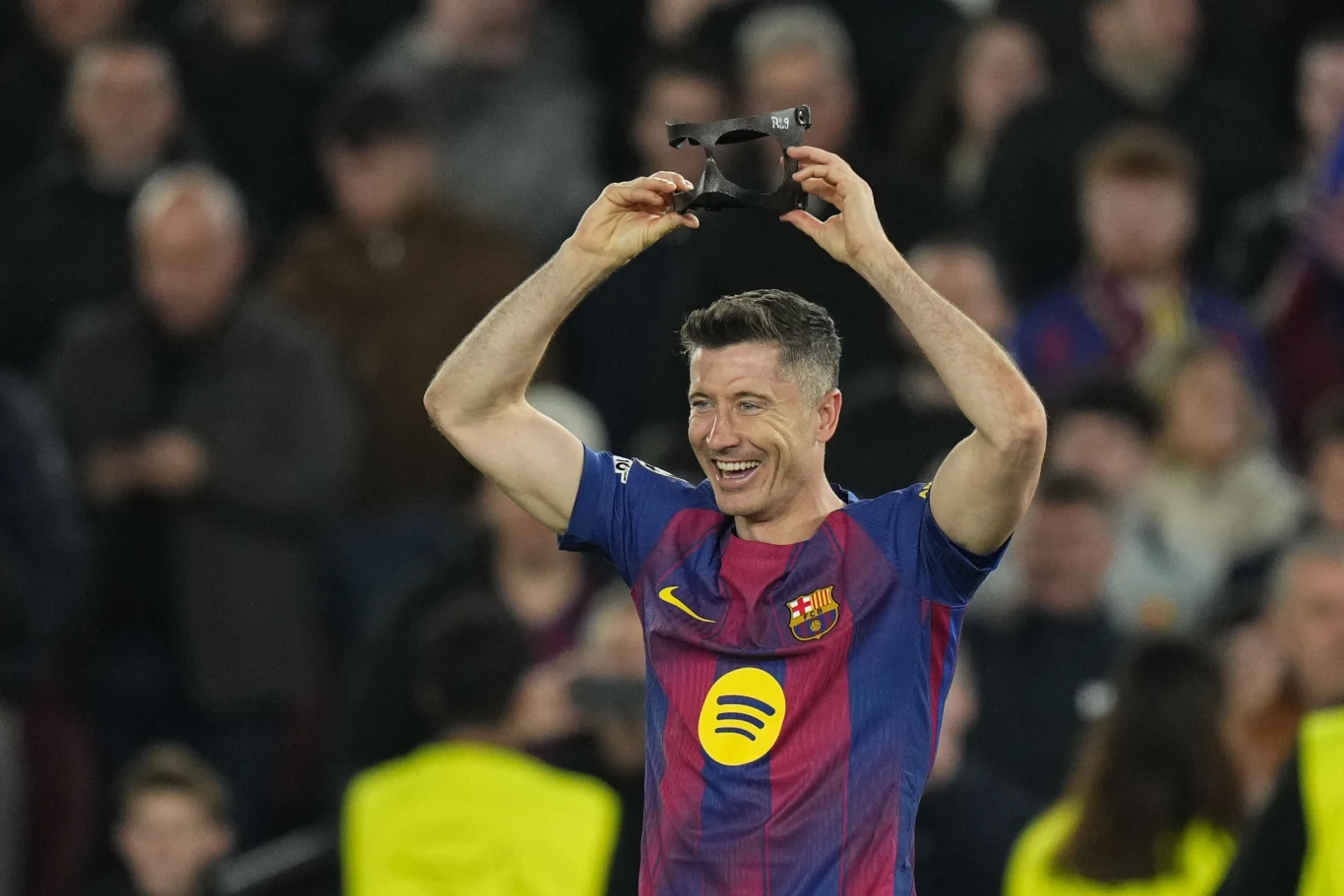piłkarz w koszulce FC Barcelony uśmiecha się i trzyma na głowie piłkarską maskę ochronną; w tle rozmyty tłum kibiców