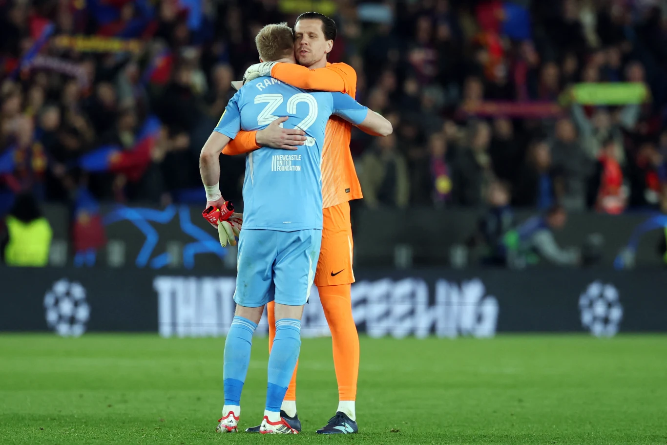 Dwóch bramkarzy w sportowych strojach obejmuje się na murawie stadionu, wokół widoczni są kibice na trybunach i charakterystyczne elementy oprawy meczu piłkarskiego.