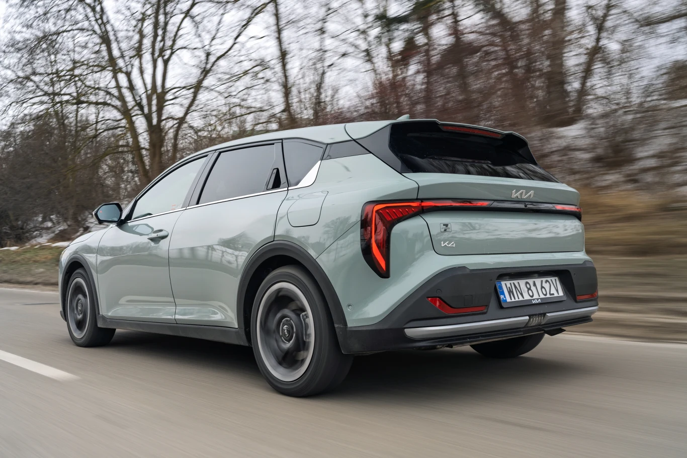 jasnozielony samochód Kia EV9 jedzie po asfaltowej drodze, nowoczesny design nadwozia podkreślają wyraziste tylne światła LED oraz kontrastujące czarne elementy karoserii, w tle bezlistne drzewa i zimowy krajobraz