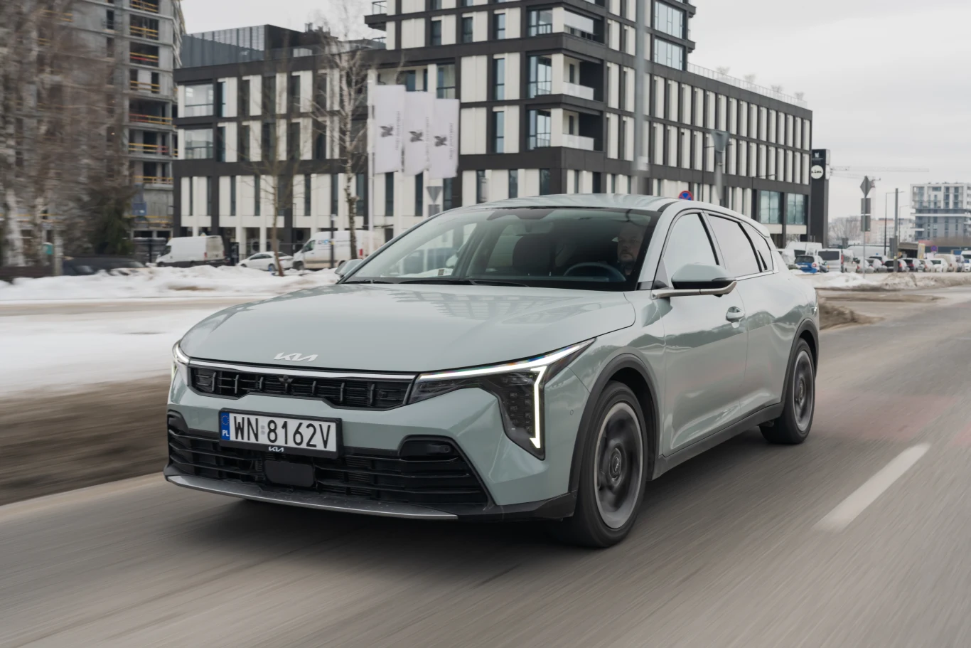 nowoczesny samochód osobowy Kia o jasnoszarym nadwoziu porusza się po miejskiej ulicy, w tle widoczne wielopiętrowe budynki i gołe drzewa, panuje zimowa aura