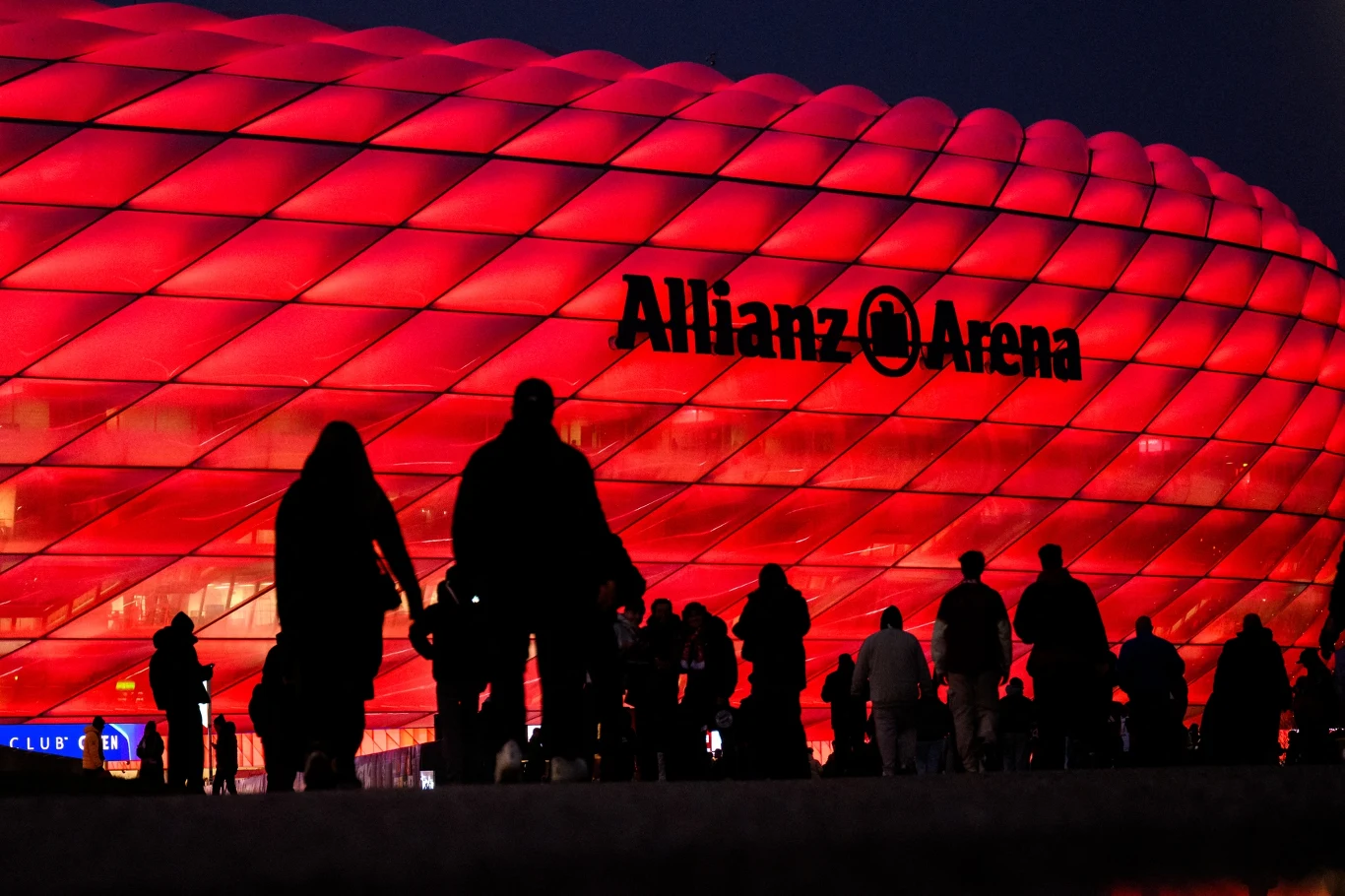 Tłum ludzi idący w kierunku jasno oświetlonego, czerwonego stadionu Allianz Arena nocą.