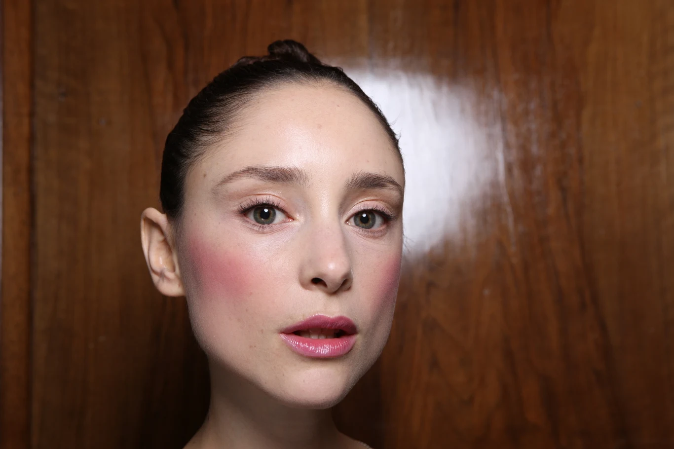 kobieta o jasnej karnacji z różowym makijażem policzków i ust, lekko rozchylone usta, włosy związane gładko do tyłu, drewniane tło
