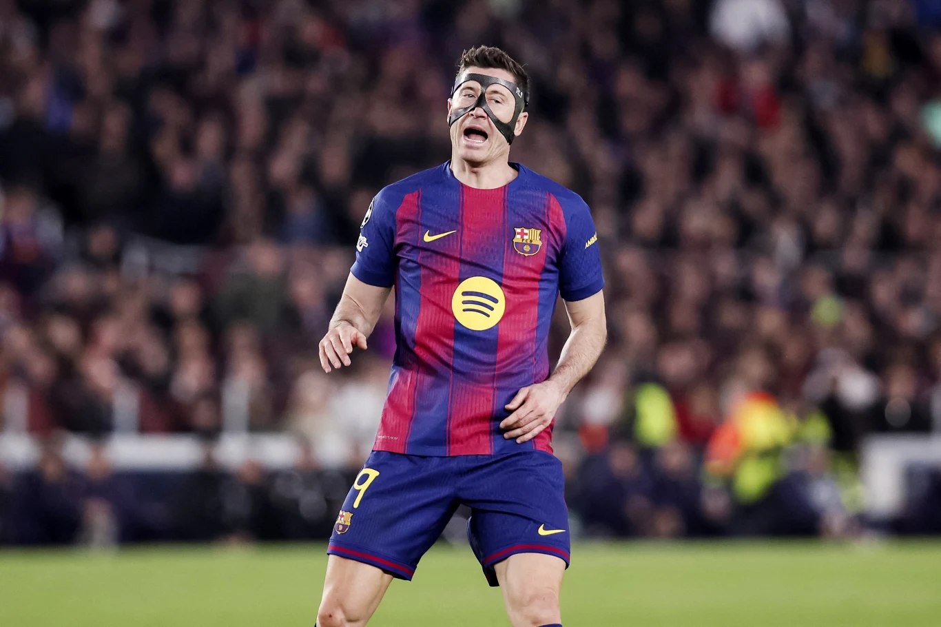 piłkarz w granatowo-bordowej koszulce FC Barcelony z numerem 9 i czarną maską ochronną na twarzy wyraża silne emocje na tle rozmytego stadionu pełnego kibiców