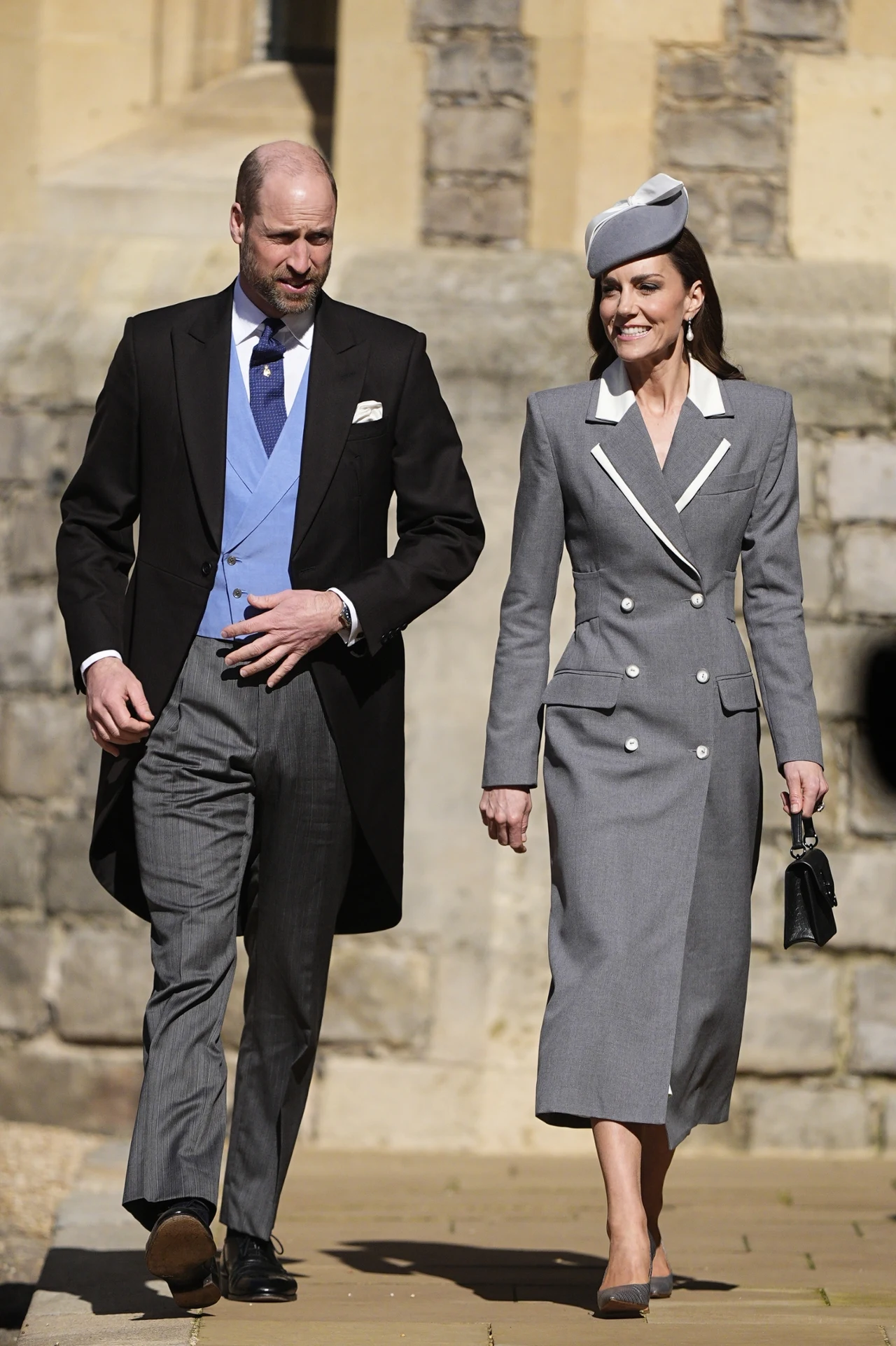 Elegancko ubrany mężczyzna w garniturze z frakiem i jasnoniebieską kamizelką idzie obok kobiety w szarym, dopasowanym płaszczu z białymi akcentami, trzymającej czarną torebkę i noszącej stylowy, szary kapelusz; oboje wyglądają na uśmiechniętych i są na...