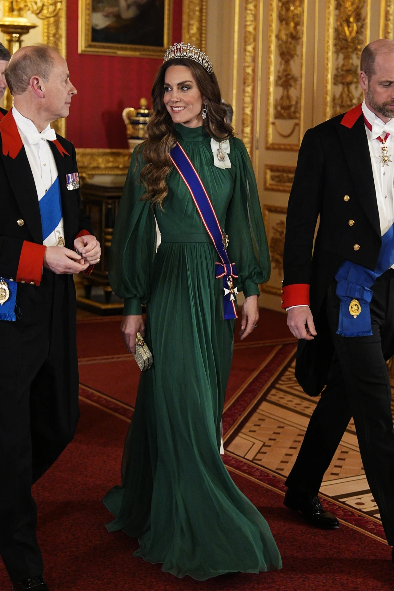 Kobieta w zielonej, długiej, eleganckiej sukni z tiarą na głowie spaceruje po pałacowym wnętrzu obok dwóch mężczyzn w formalnych strojach z orderami; wszyscy otoczeni są bogato zdobionymi, złotymi dekoracjami ściennymi.