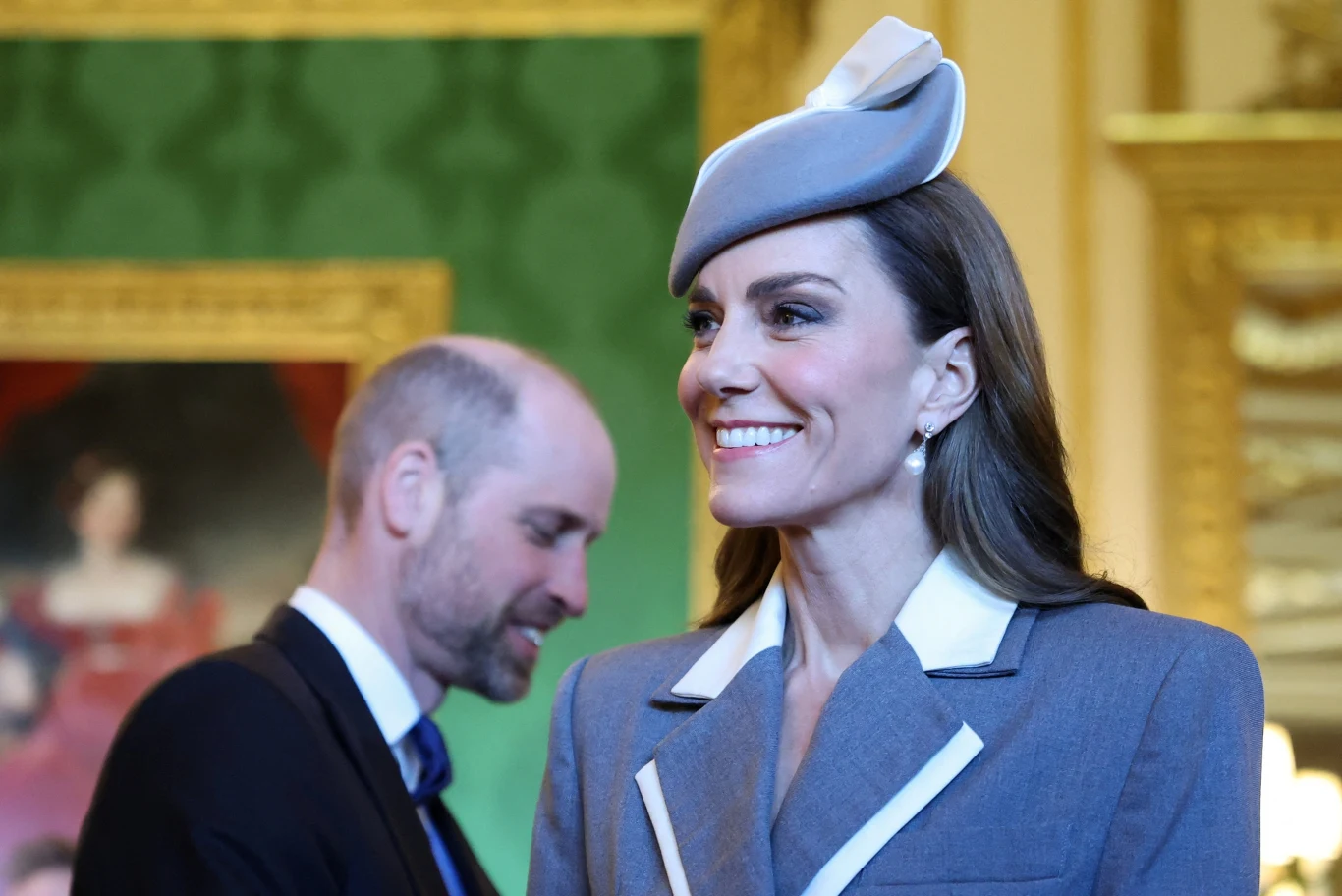Uśmiechnięta kobieta w eleganckim szarym kostiumie z białymi akcentami i ozdobnym kapeluszem, stojąca na tle dekoracyjnych, złoconych ram obrazów w stylowym wnętrzu; w tle mężczyzna w garniturze z lekko pochyloną głową.