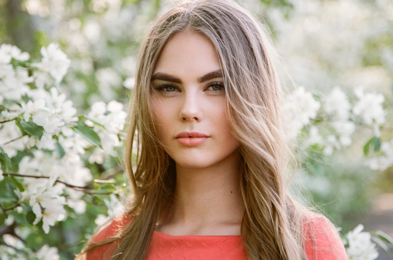 młoda kobieta o długich, jasnych włosach i subtelnym makijażu, ubrana w koralową sukienkę, stoi wśród kwitnących gałęzi drzewa owocowego, patrząc prosto w obiektyw