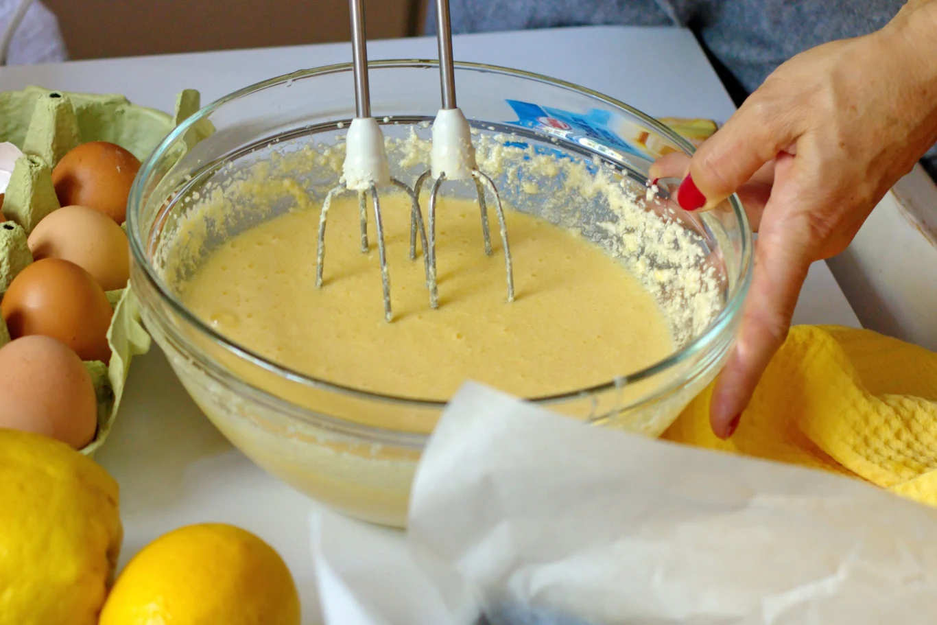szklana miska z ubijaną masą ciasta mikserem ręcznym, obok jajka i cytryny, dłoń trzymająca miskę