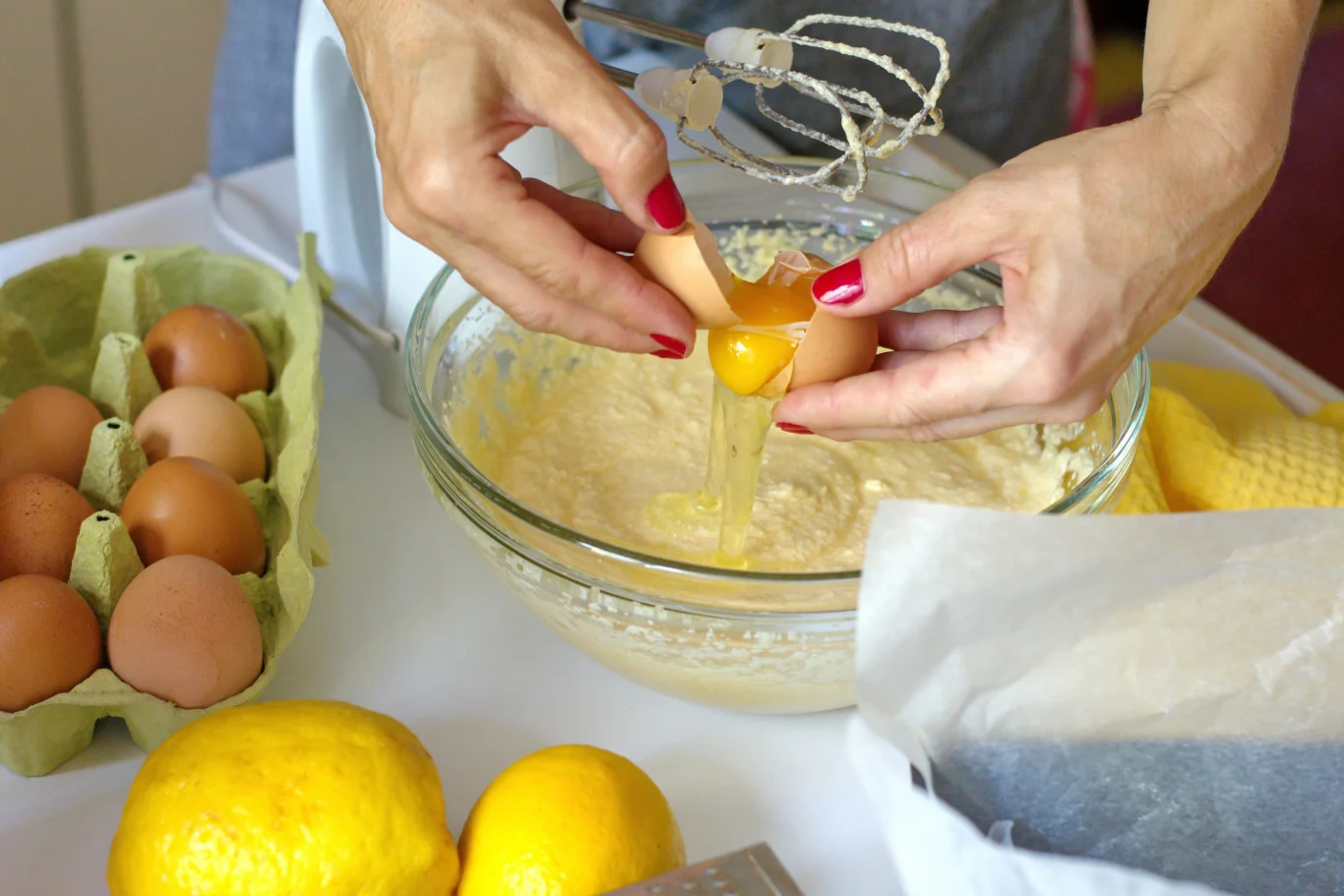 Ręce rozbijające jajko nad szklaną miską z ciastem, obok mikser, tacka z jajkami, cytryny i tarka.
