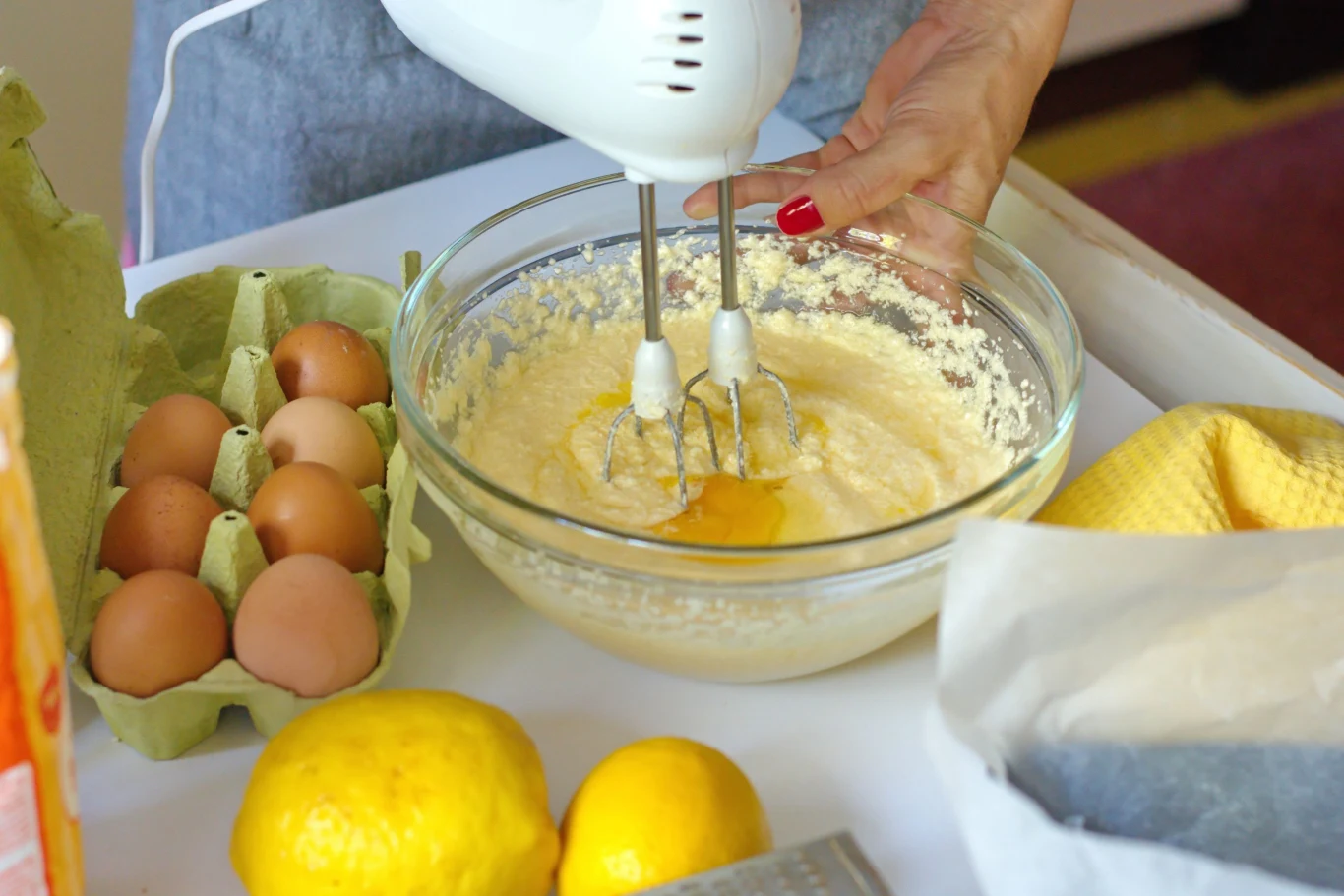dłoń trzymająca mikser elektryczny ubijający ciasto w szklanej misce, obok leżą jajka w wytłaczance, dwie cytryny oraz tarka