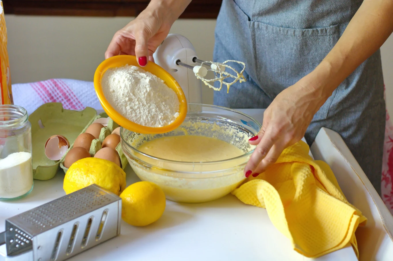 osoba w szarej kuchennej fartuchu wsypuje mąkę do miski z ciastem, obok znajdują się cytryny, jajka, tarka, ręcznik kuchenny i mikser