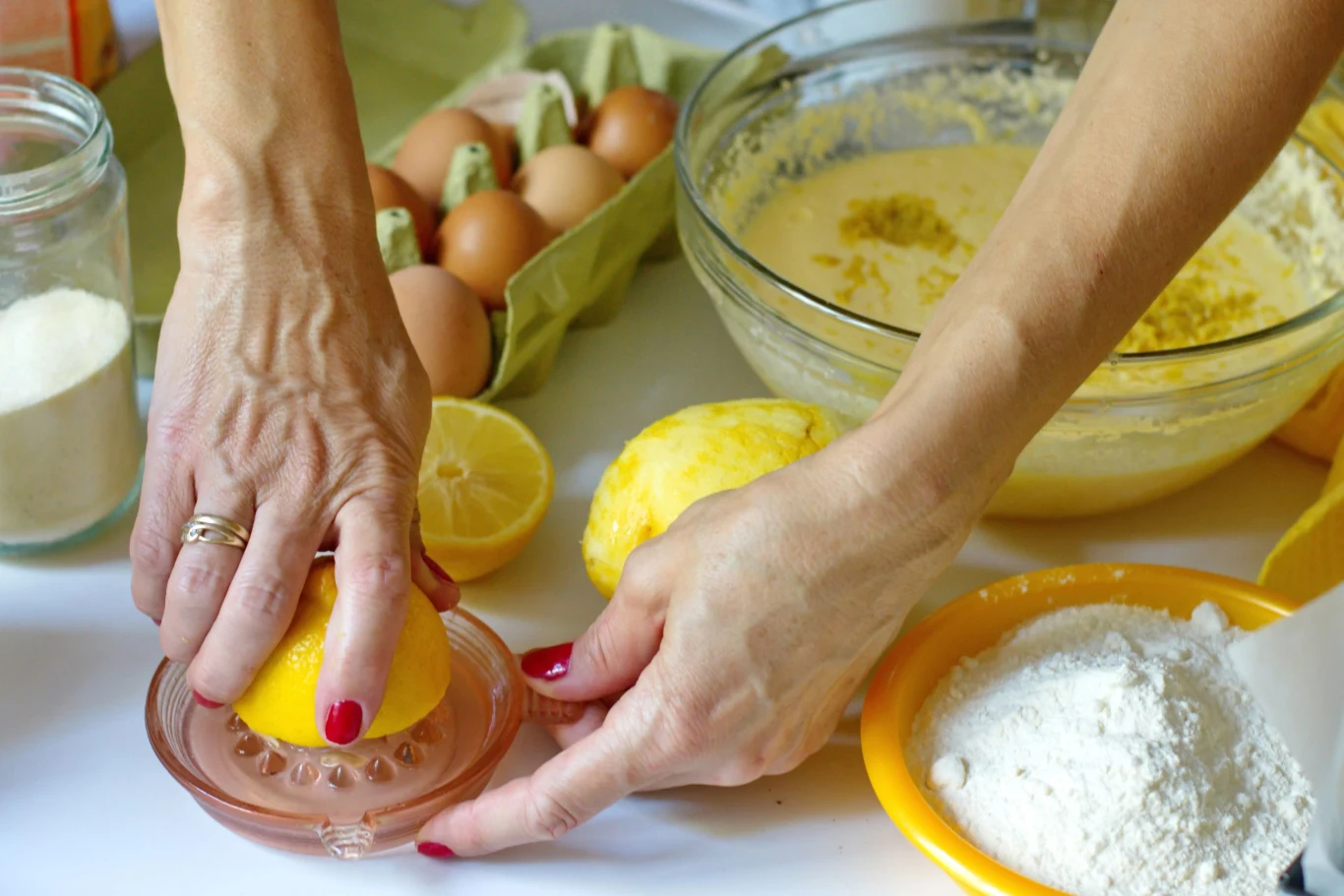 wyciskanie soku z cytryny do szklanej wyciskarki, w tle miska z ciastem, talerz z mąką, kilka jajek oraz przekrojona cytryna