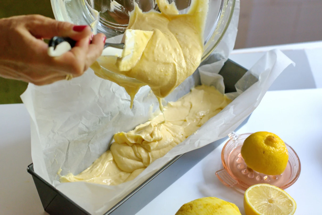 Gęste ciasto cytrynowe wylewane do prostokątnej formy wyłożonej papierem do pieczenia, obok leżą przekrojona cytryna, wyciskacz do cytryn i cała cytryna.