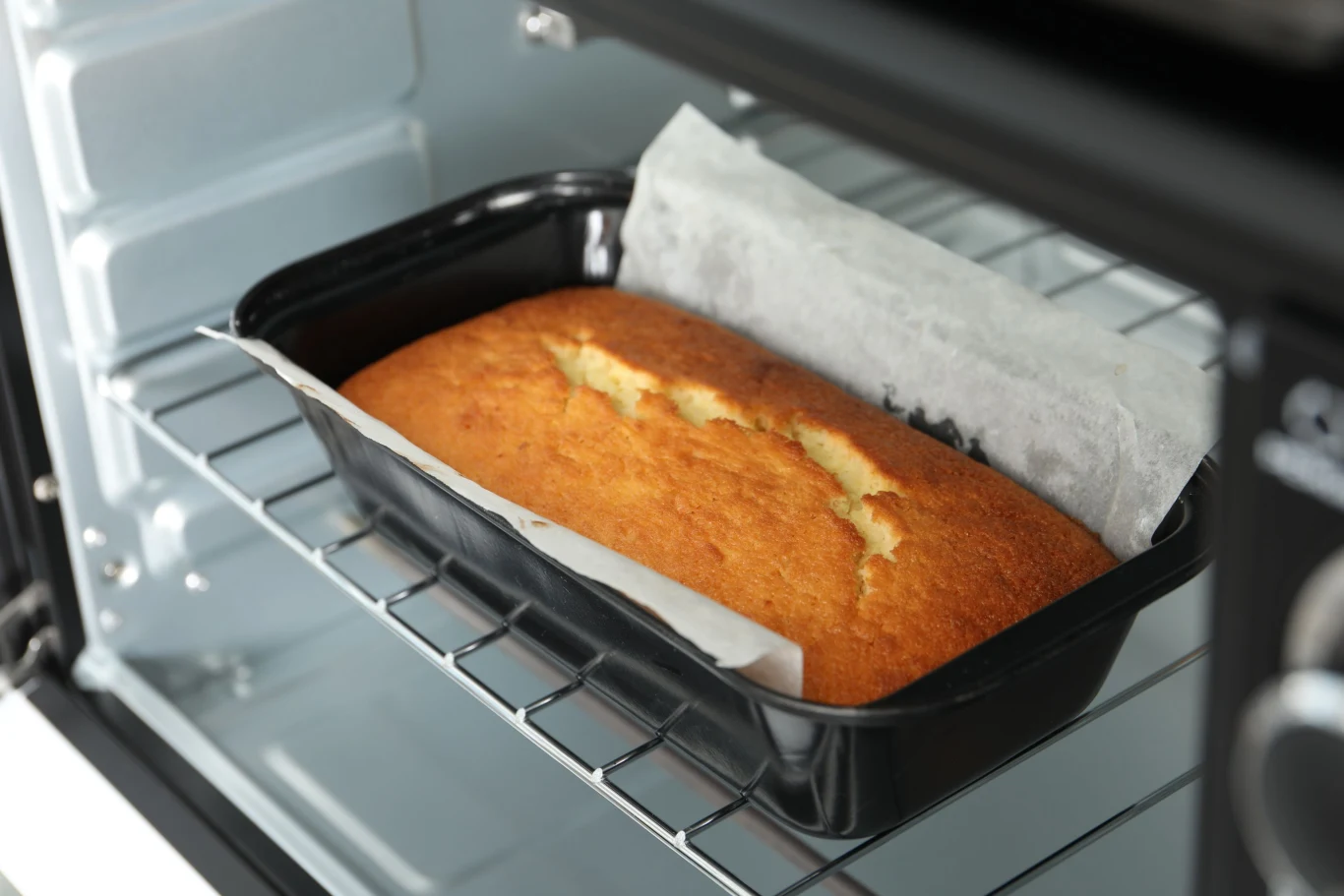 Upieczone ciasto o złotobrązowej powierzchni w czarnej, prostokątnej formie wyłożonej papierem do pieczenia, ustawione na środkowej półce otwartego piekarnika.