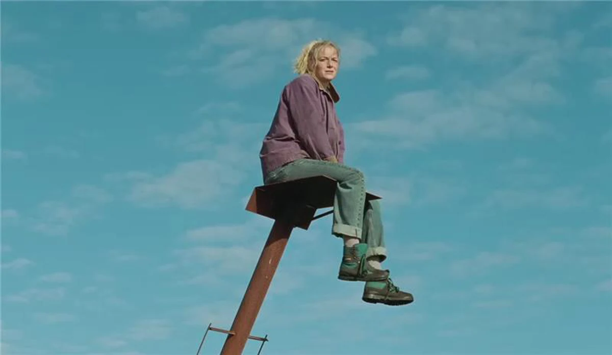 Kadr z filmu "Miłość, która zostaje" Kobieta w fioletowej kurtce i zielonych spodniach siedzi na metalowej konstrukcji, w tle niebo