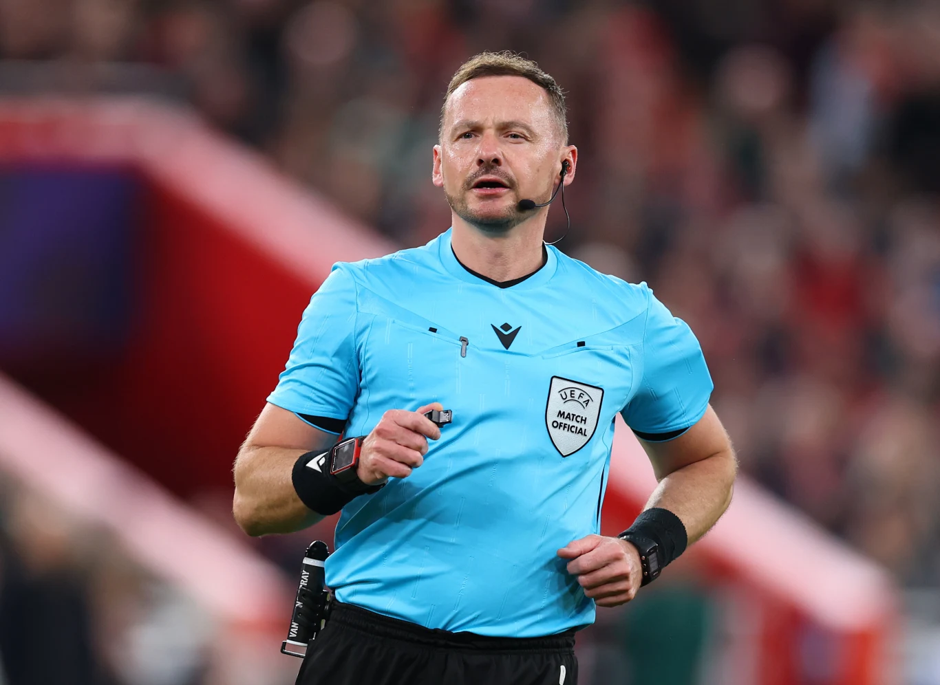 sędzia piłkarski w niebieskiej koszulce UEFA, z gwizdkiem i słuchawką na uchu, skoncentrowany podczas prowadzenia meczu