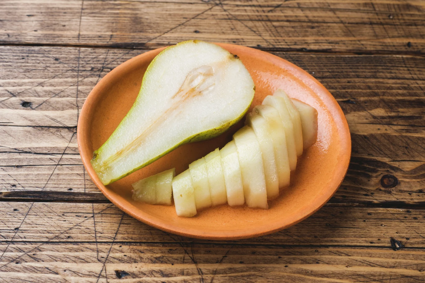 Łatwo dostępny owoc pełen wartości odżywczych. Dlaczego warto włączyć gruszki do codziennej diety? Połówka gruszki i plasterki tego owocu na pomarańczowym talerzu, na drewnianym stole.