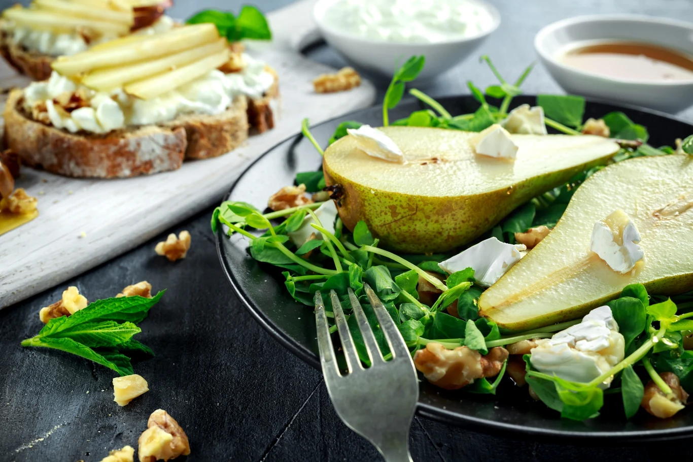 Gruszki smakowicie komponują się w wersji wytrawnej z serem i orzechami. Sałatka z rukolą, połówkami gruszki, kawałkami białego sera pleśniowego i orzechami włoskimi na czarnym talerzu, w tle chrupiące grzanki z serem, plasterkami gruszki i orzechami.