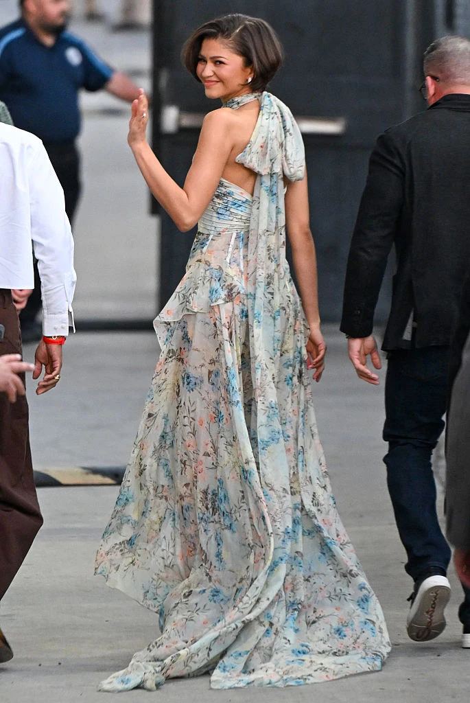 kobieta ubrana w długą, zwiewną suknię o kwiecistym wzorze, uśmiecha się i macha ręką, idąc w otoczeniu kilku osób; jej krótkie, elegancko ułożone włosy oraz delikatny makijaż podkreślają stylizację