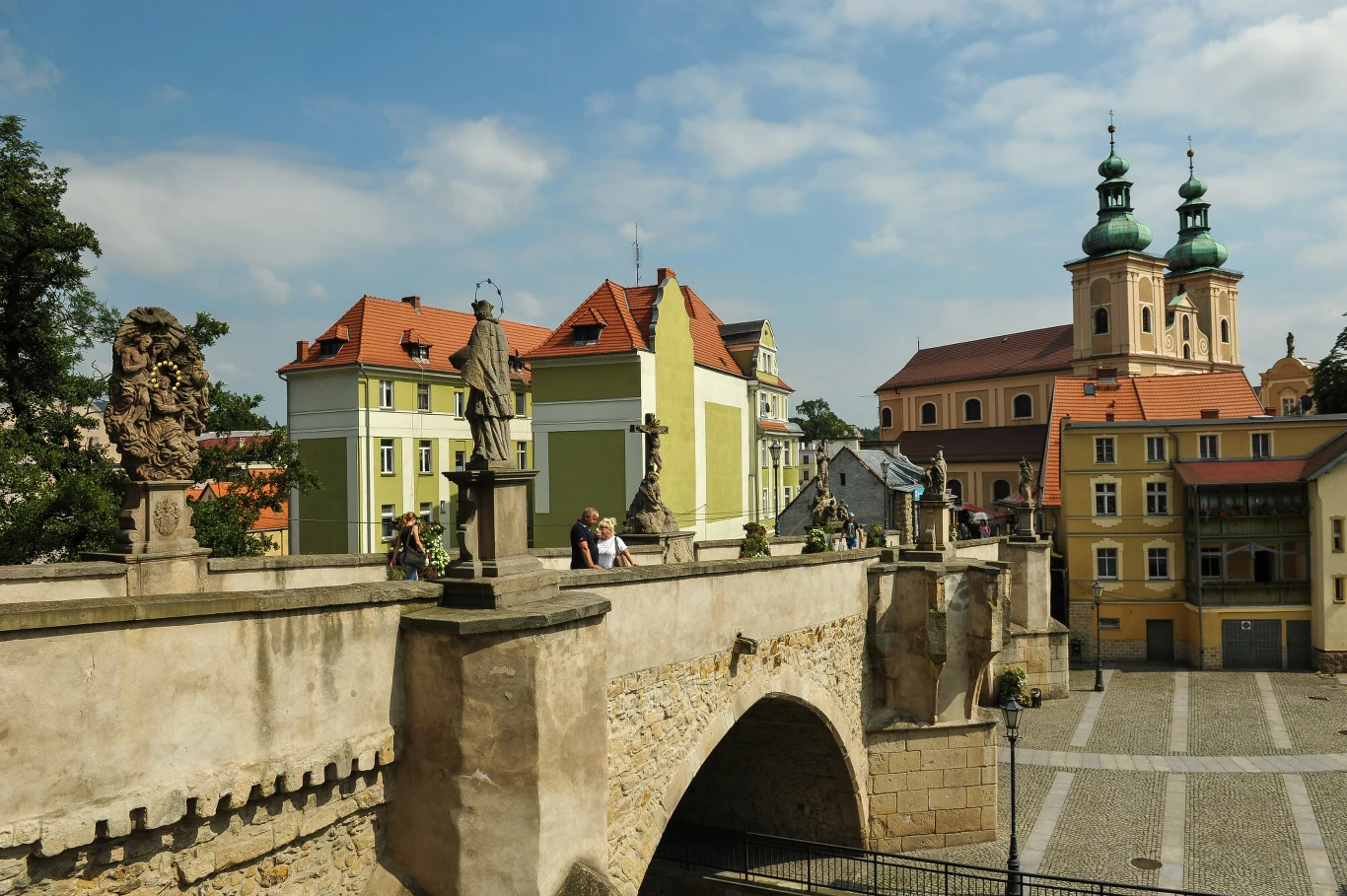 kamienny most ozdobiony barokowymi rzeźbami, prowadzi do placu otoczonego kolorowymi kamienicami oraz dużym kościołem o dwóch zielonych wieżach