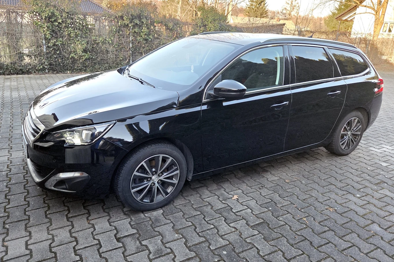 czarny samochód kombi zaparkowany na brukowanym podjeździe, widoczny z boku i częściowo z przodu, auto jest czyste i zadbane, ma przyciemnione szyby oraz nowoczesne, wieloramienne felgi