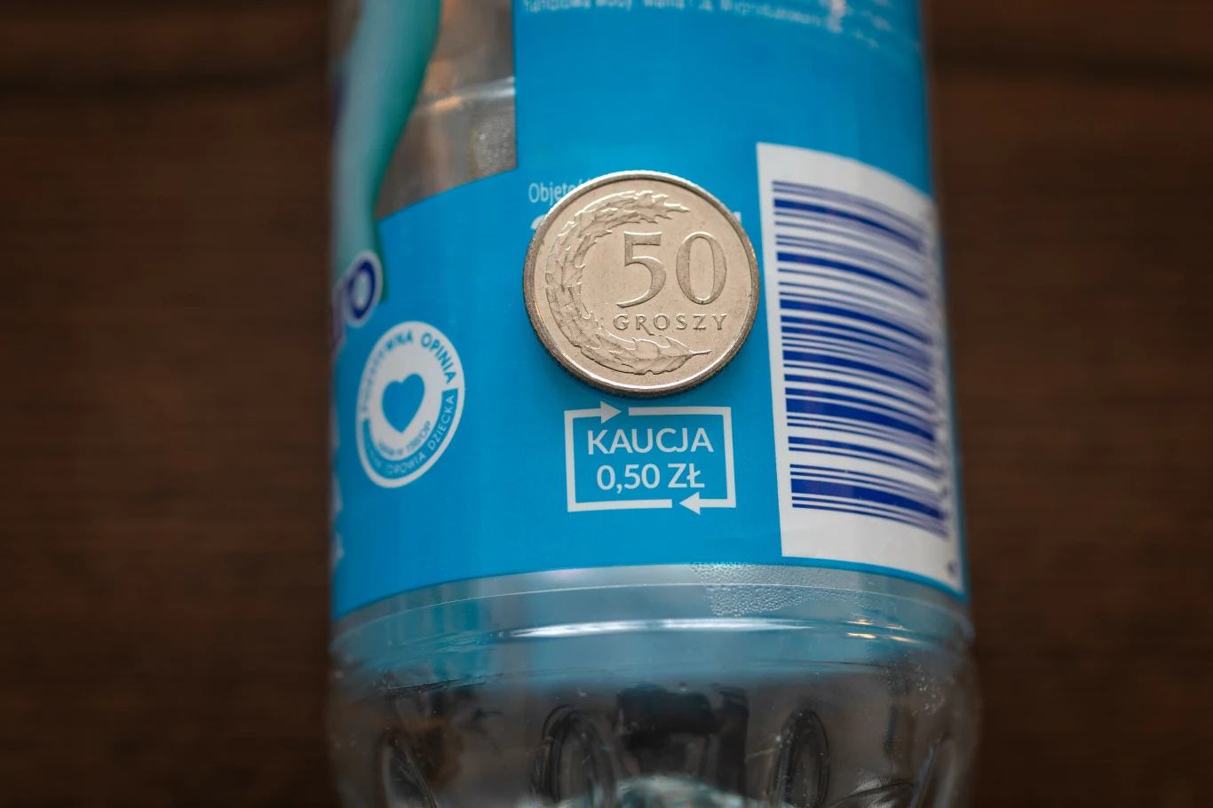 Moneta o nominale 50 groszy umieszczona na etykiecie plastikowej butelki z informacją o kaucji 0,50 zł, obok widoczny kod kreskowy na niebieskim tle.