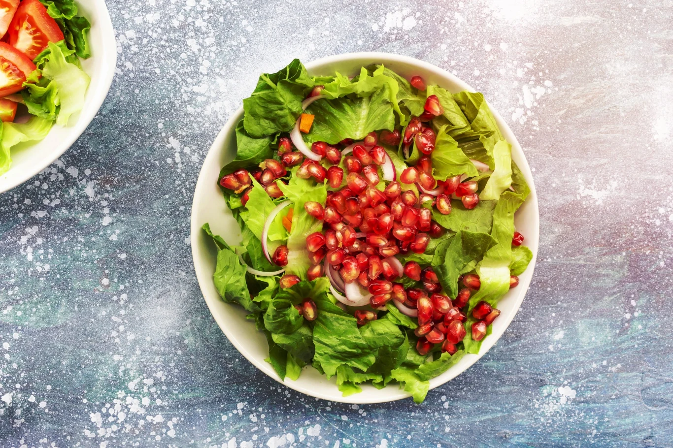 sałatka z liści sałaty, plasterków czerwonej cebuli i dużą ilością ziaren granatu podana w białej misce na niebiesko-białym tle