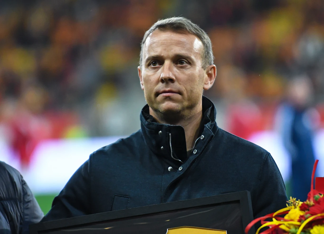 mężczyzna w ciemnej kurtce stoi na tle rozmytego stadionu, trzymając w dłoniach tablicę i bukiet czerwono-żółtych kwiatów